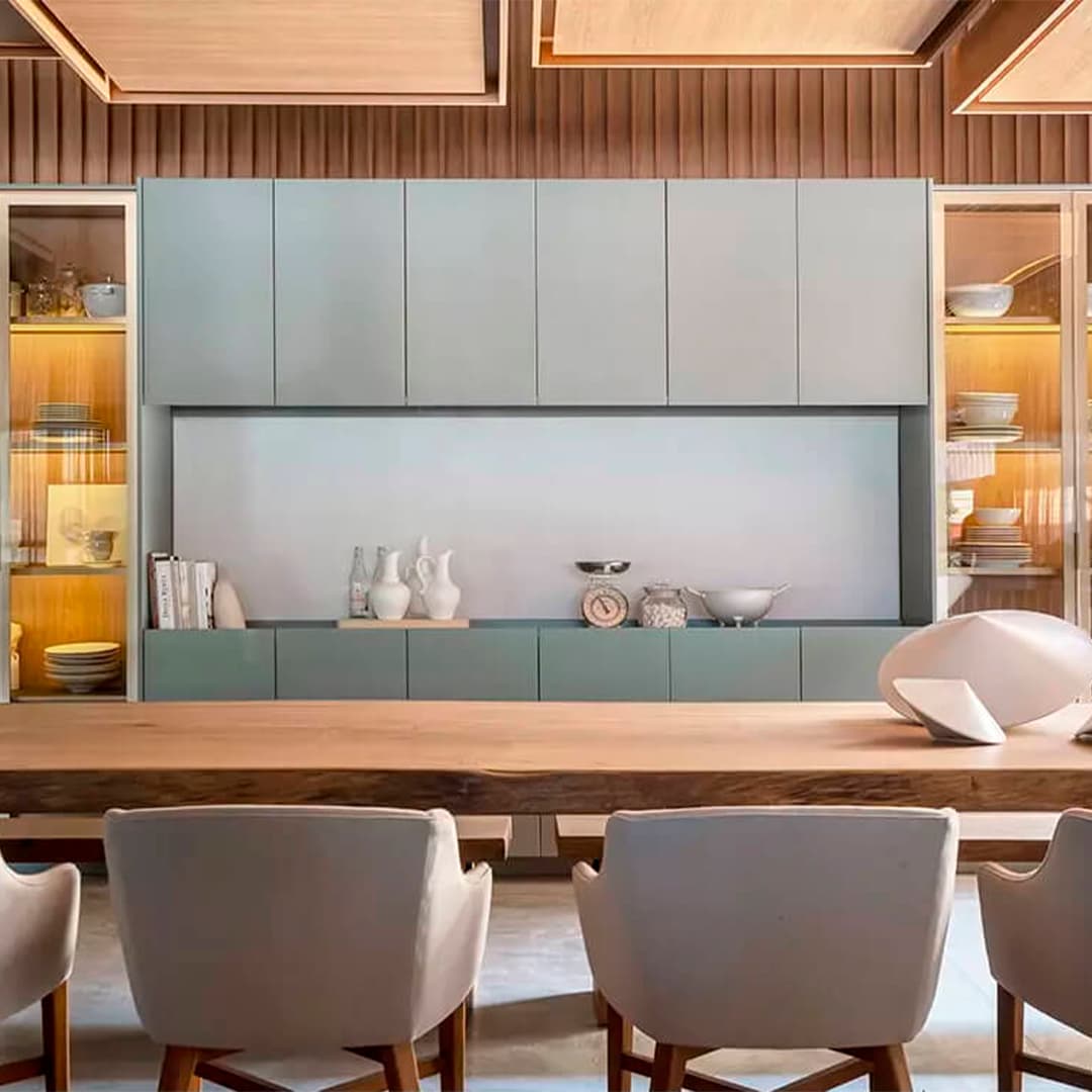 Sala de jantar com marcenaria verde sálvia, cristaleiras iluminadas e mesa em madeira viva.